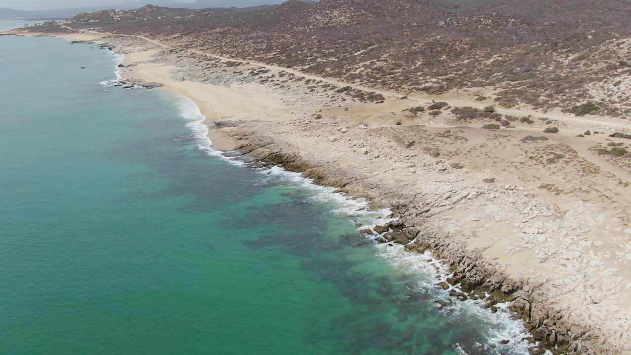 Mid-clip aerial frame at Punta Gorda Beach, with a broad turquoise ocean area, white waves hitting rocks, sandy flats, dry scrub hills, and no visible people.