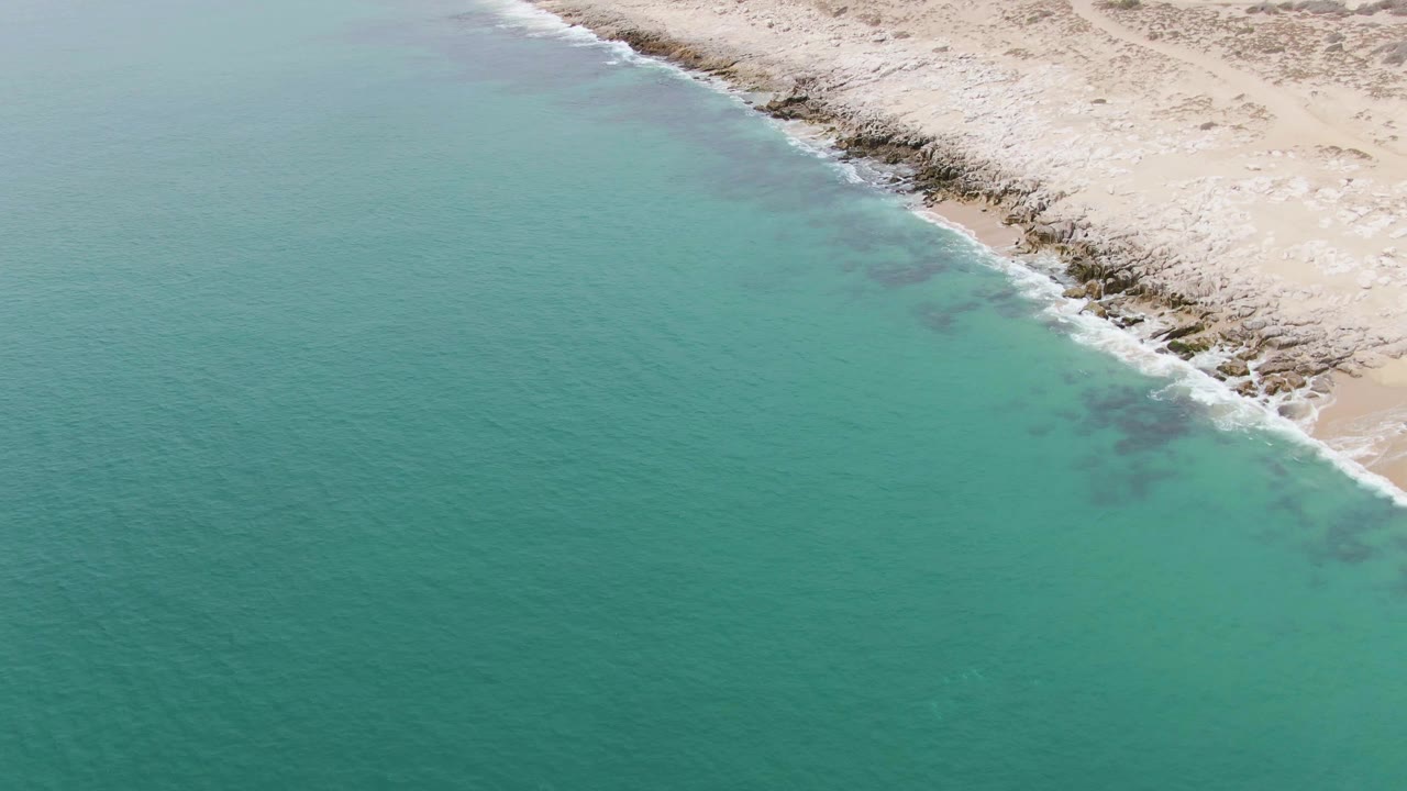 Aerial view of Punta Gorda Beach with calm turquoise water, a thin white surf line, pale sand, exposed rocks, and no visible people.