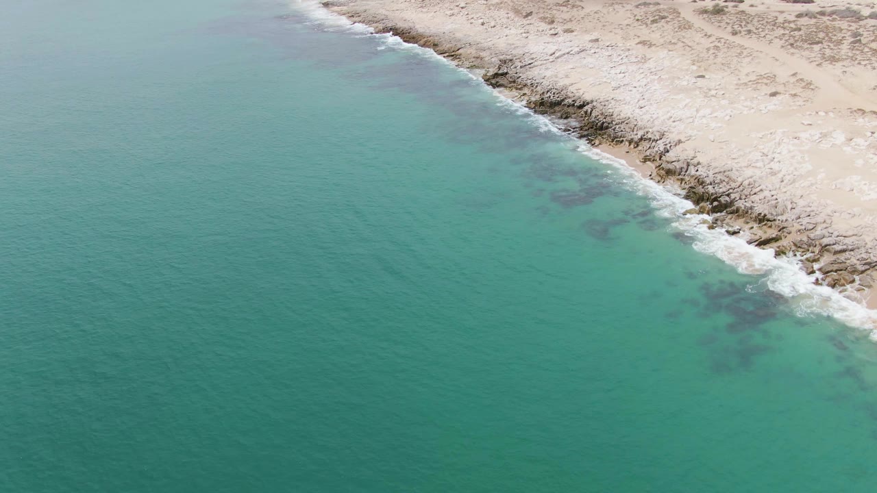 Aerial frame at Punta Gorda Beach showing broad turquoise water with darker underwater patches near the rocky shoreline and no visible people.