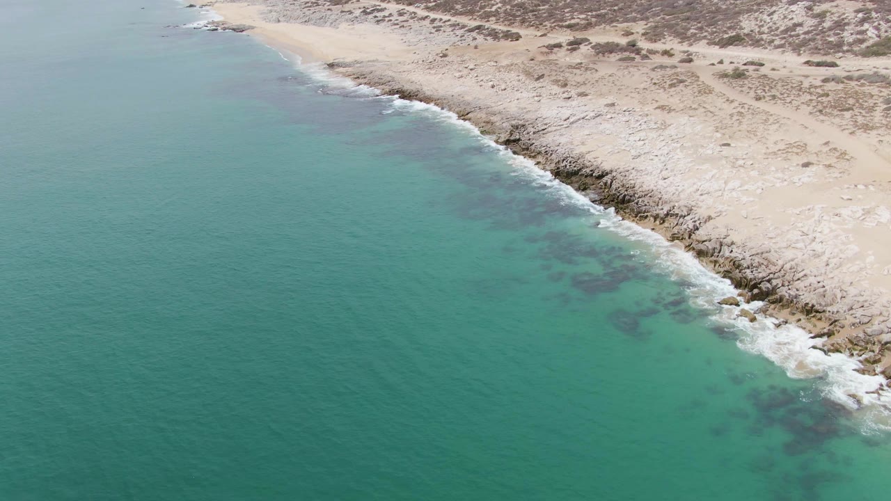 Punta Gorda Beach seen from above, with the coastline running diagonally through the frame, turquoise water on the left, pale rocky beach on the right, and no visible people.