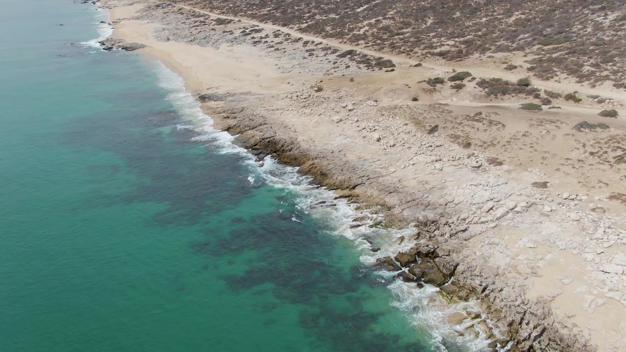 Aerial view at Punta Gorda Beach, Baja California Sur, with turquoise water on the left, pale rocky shore on the right, sandy dunes inland, and no visible people.