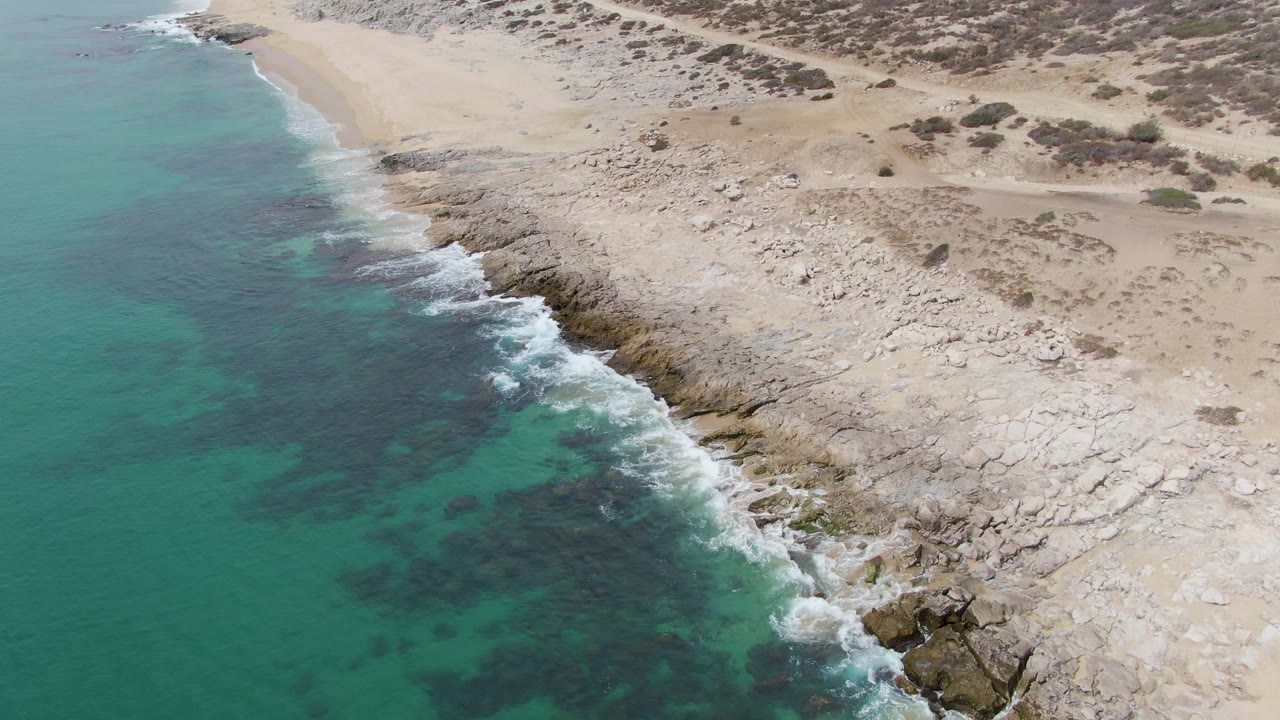 Aerial view at Punta Gorda Beach with clear turquoise water, a diagonal band of surf on the rocks, pale sand and scrub-covered dunes inland, and no visible people.