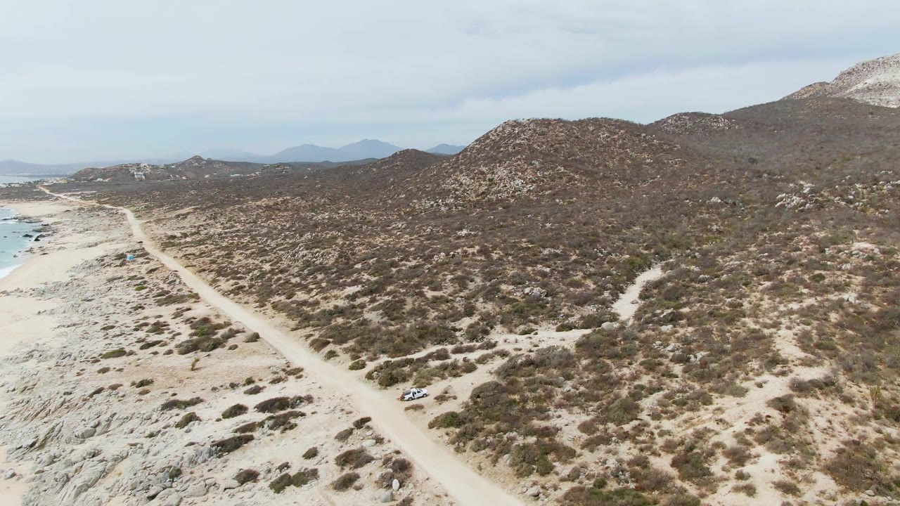 Aerial frame at Punta Gorda Beach, Baja California Sur, with the dirt road running from the lower beach area toward distant hills, ocean water at left, scrubland across the slope, and no visible people.