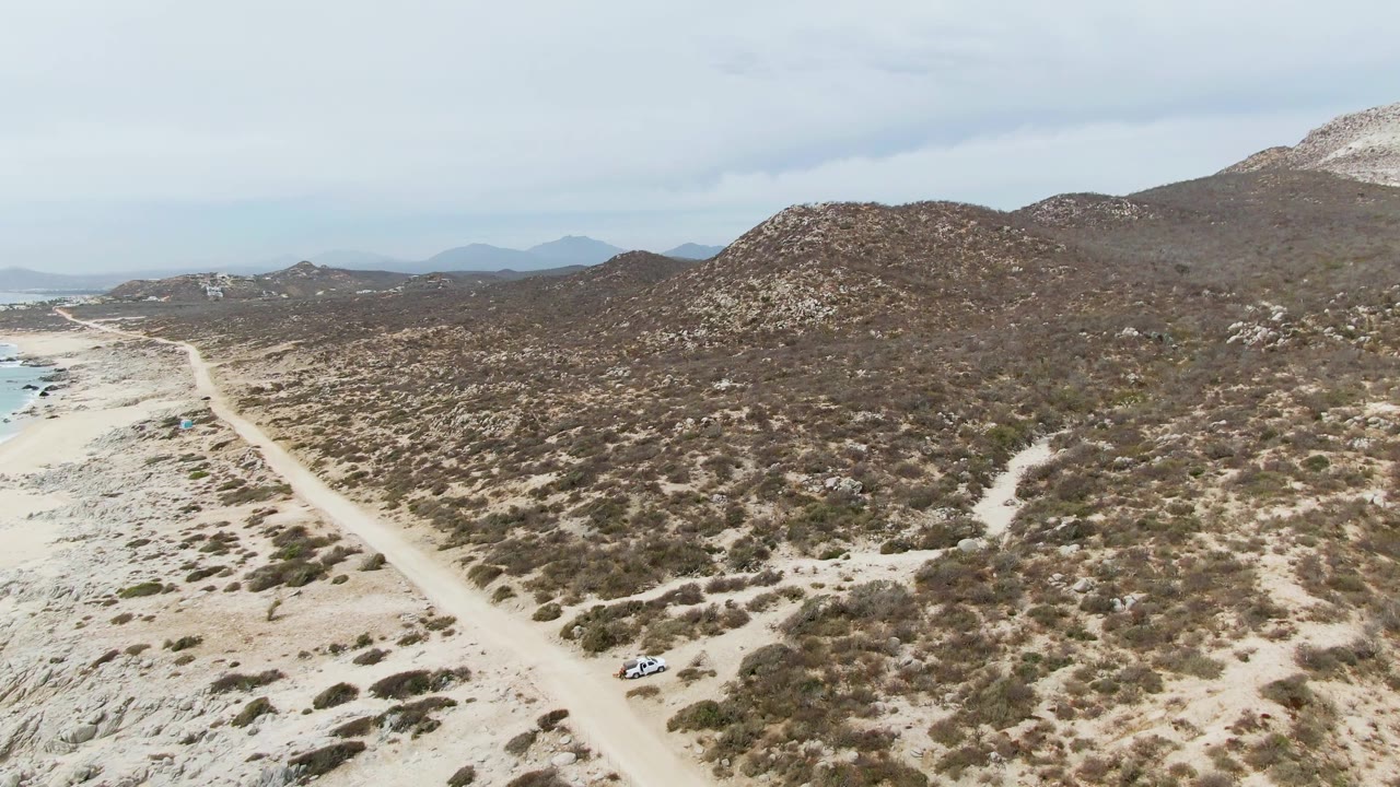 Aerial frame at Punta Gorda Beach, Baja California Sur, showing the road set between pale beach terrain and brown-green scrub hills, with blue ocean at the far left and no visible people.