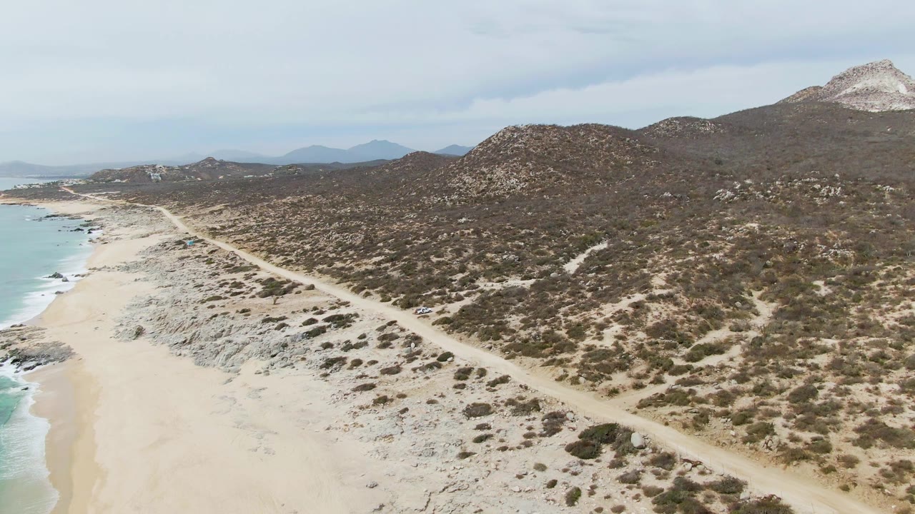 Aerial frame at Punta Gorda Beach shows a pale dirt road running beside sand, turquoise surf, dry hills, distant mountains, cloudy sky, and no visible people.