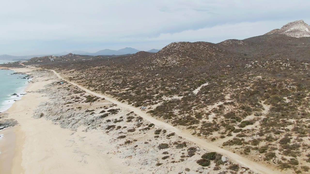 Coastal dirt road curves along sand with hills and ocean visible.