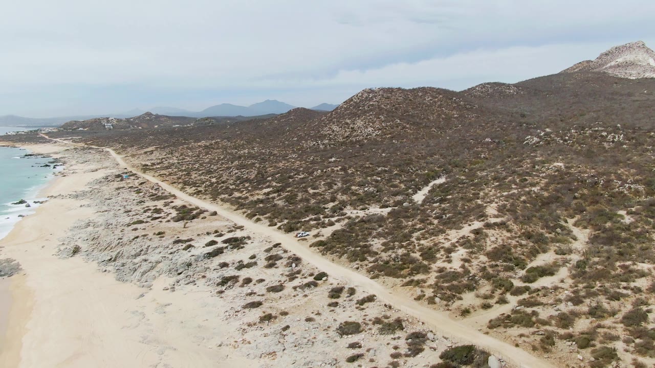 At Punta Gorda Beach, a small white vehicle is visible on the dirt road near the right side of the road line, with scrub hills, sandy beach, ocean, and no visible people.