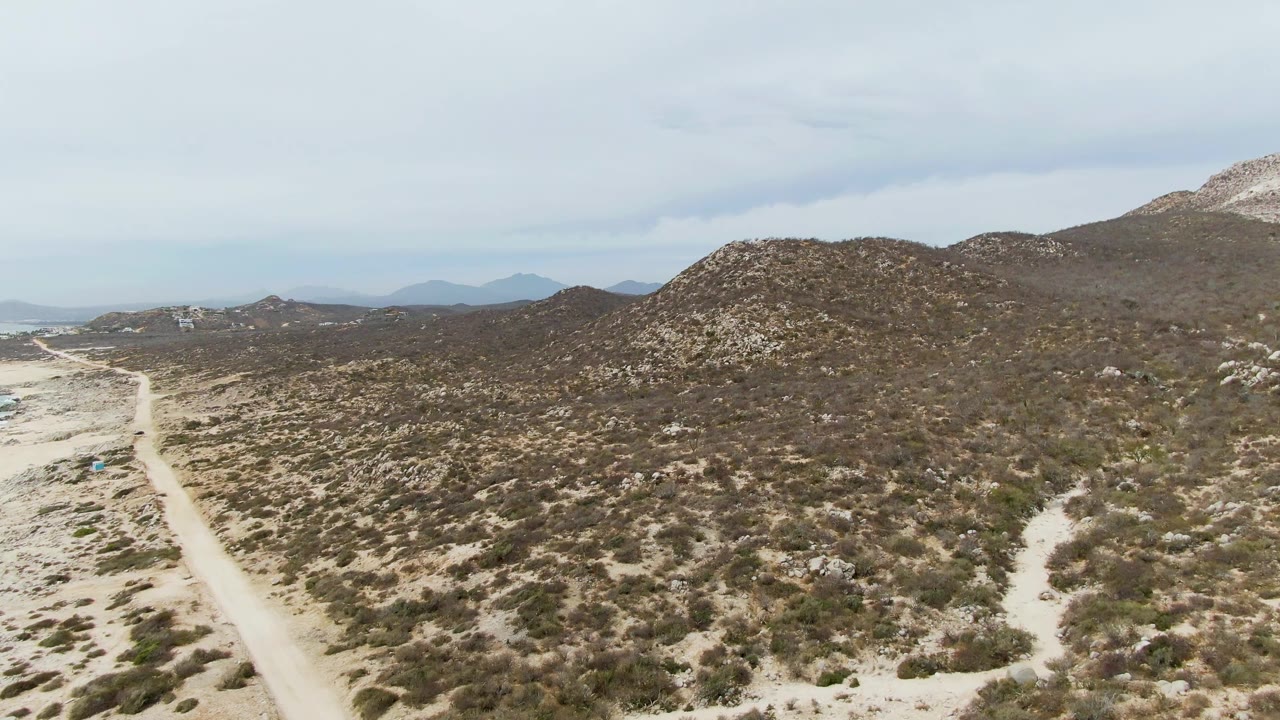 Aerial frame from Punta Gorda Beach, Baja California Sur showing a pale dirt road along scrub-covered sandy terrain, rocky hills, distant hazy ridges, and no visible people.