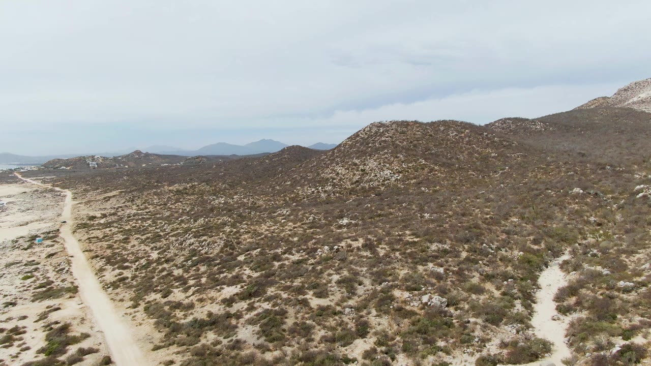 Aerial frame from Punta Gorda Beach, Baja California Sur with the dirt road running up the left side, scrub across sandy ground, a rounded rocky hill in the center, and no visible people.