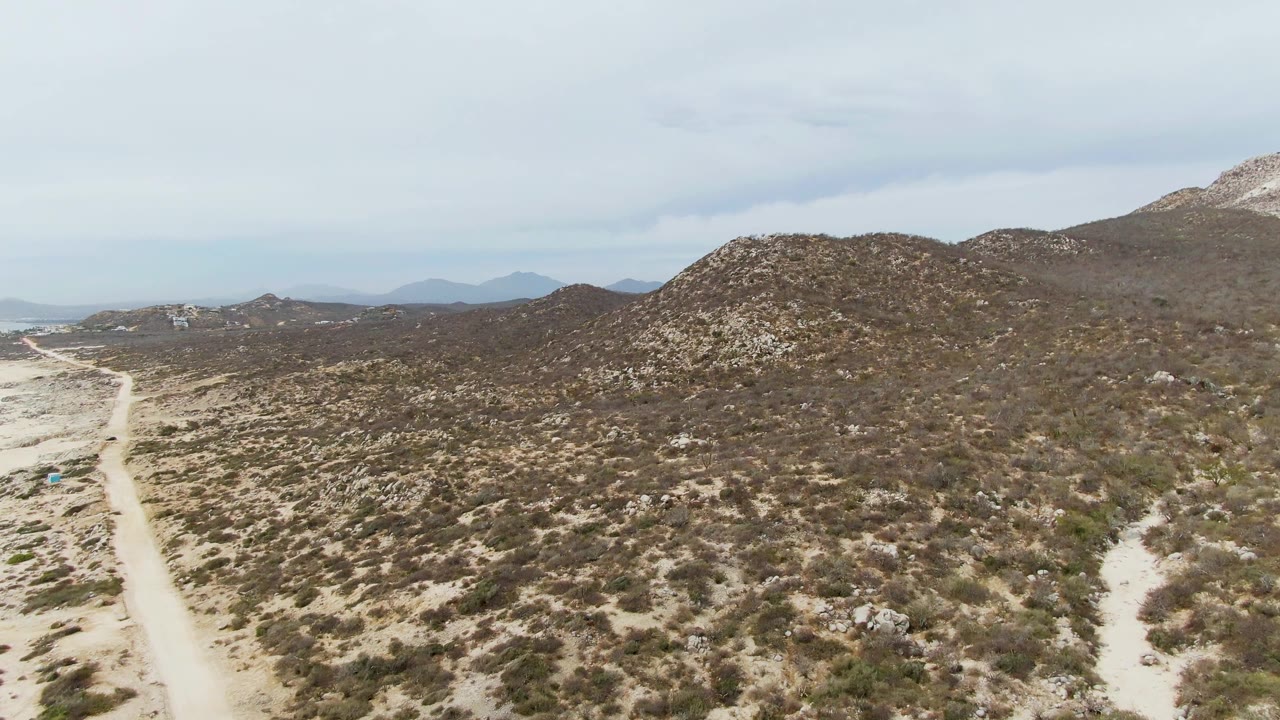 Aerial frame from Punta Gorda Beach, Baja California Sur showing pale dirt tracks on both sides of scrubby desert, low rocky slopes, muted overcast light, and no visible people.