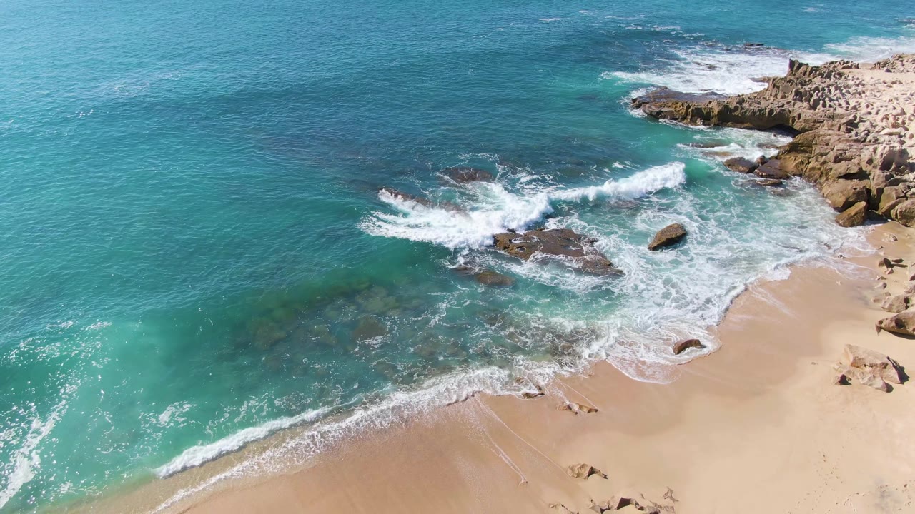 At 0:03, the aerial view shows a stronger wave crest breaking over the central rocks at Punta Gorda Beach, with open turquoise water on the left and no visible people.