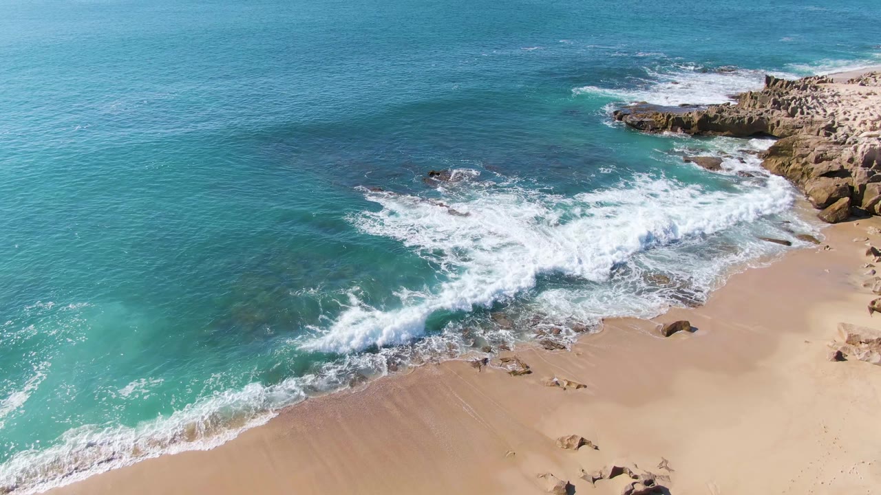 Aerial frame over Punta Gorda Beach showing turquoise water curling into white foam across dark rocks and tan sand, with no visible people.