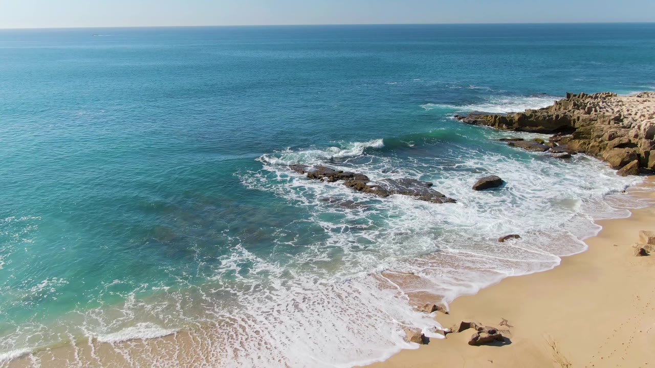 Aerial frame at Punta Gorda Beach, Baja California Sur, showing turquoise water, white foam, sandy shoreline, dark surf rocks, a rocky outcrop at right, and no visible people.