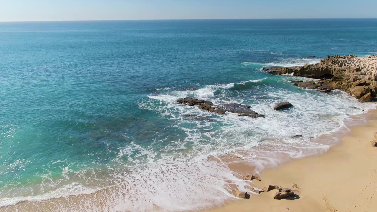 Aerial frame at Punta Gorda Beach, Baja California Sur, with a fresh band of white foam curling around midshore rocks, pale sand below, open blue ocean above, and no visible people.