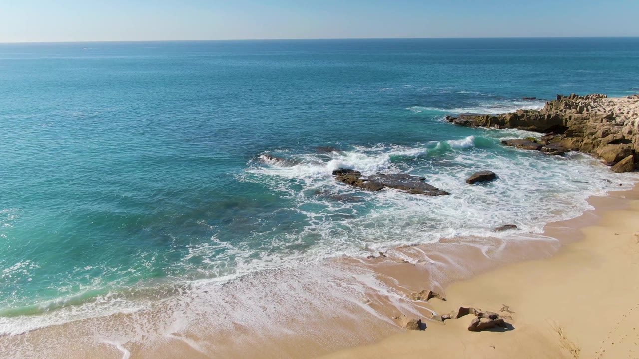 Aerial frame at Punta Gorda Beach, Baja California Sur, showing foam pushed farther across wet sand, exposed rocks near the lower shoreline, a rocky outcrop on the right, and no visible people.