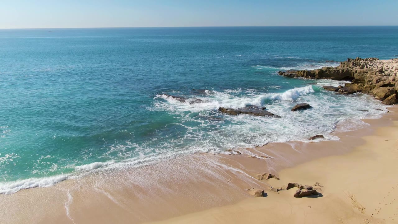 Wide aerial beach view with blue water, white surf, sand, and rocks.