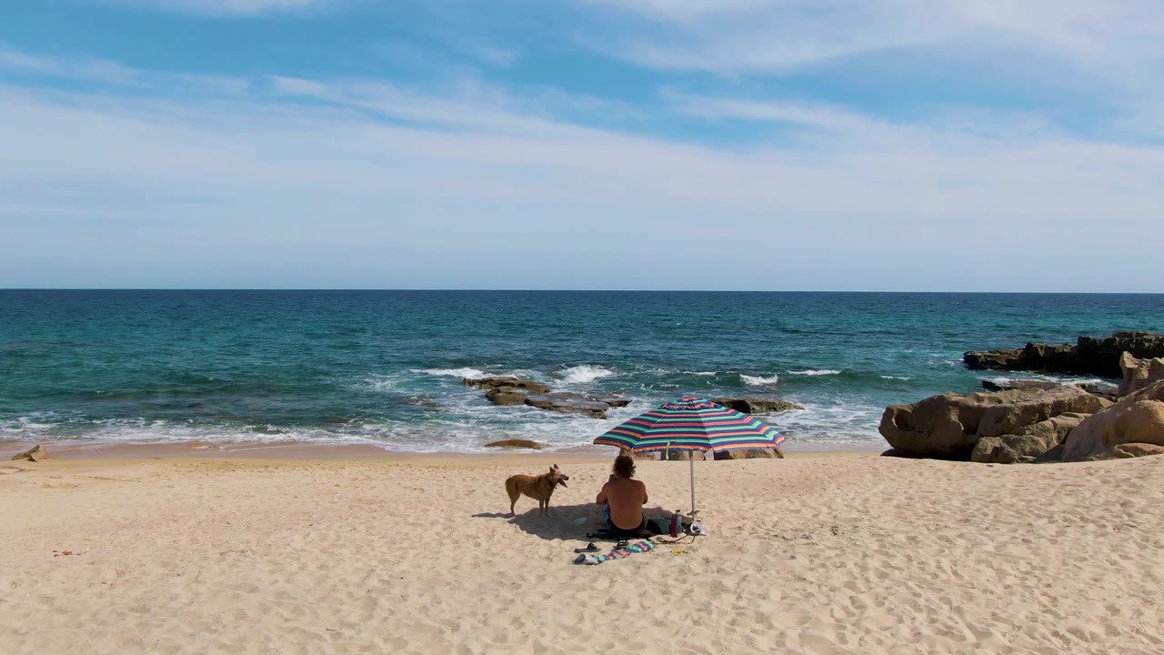 At Punta Gorda Beach in Baja California Sur, a seated beachgoer and a dog rest near a striped umbrella while small waves break behind them and dark rocks sit at the right edge.