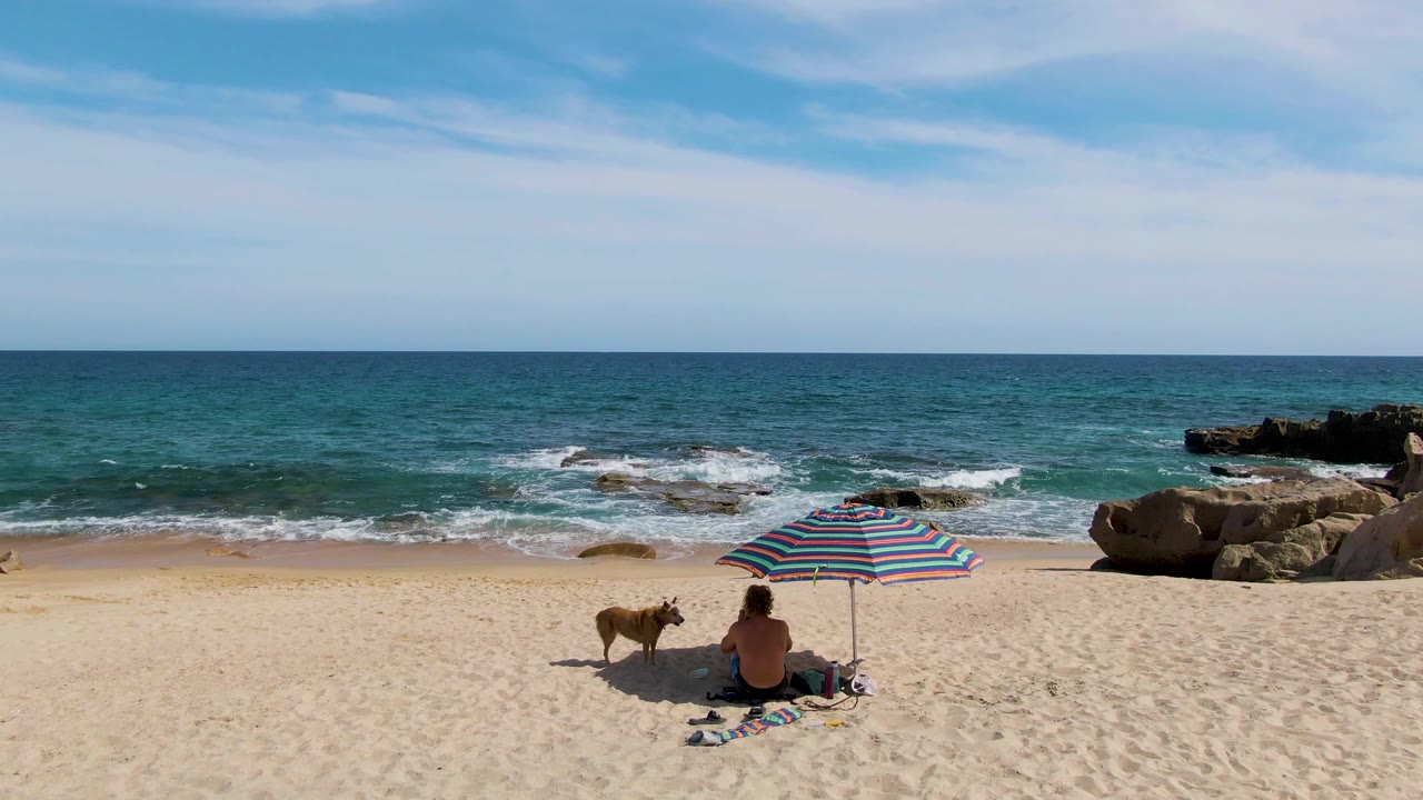 At Punta Gorda Beach in Baja California Sur, the seated person remains near the striped umbrella while the dog stands nearby and a low wave breaks around the rocks offshore.