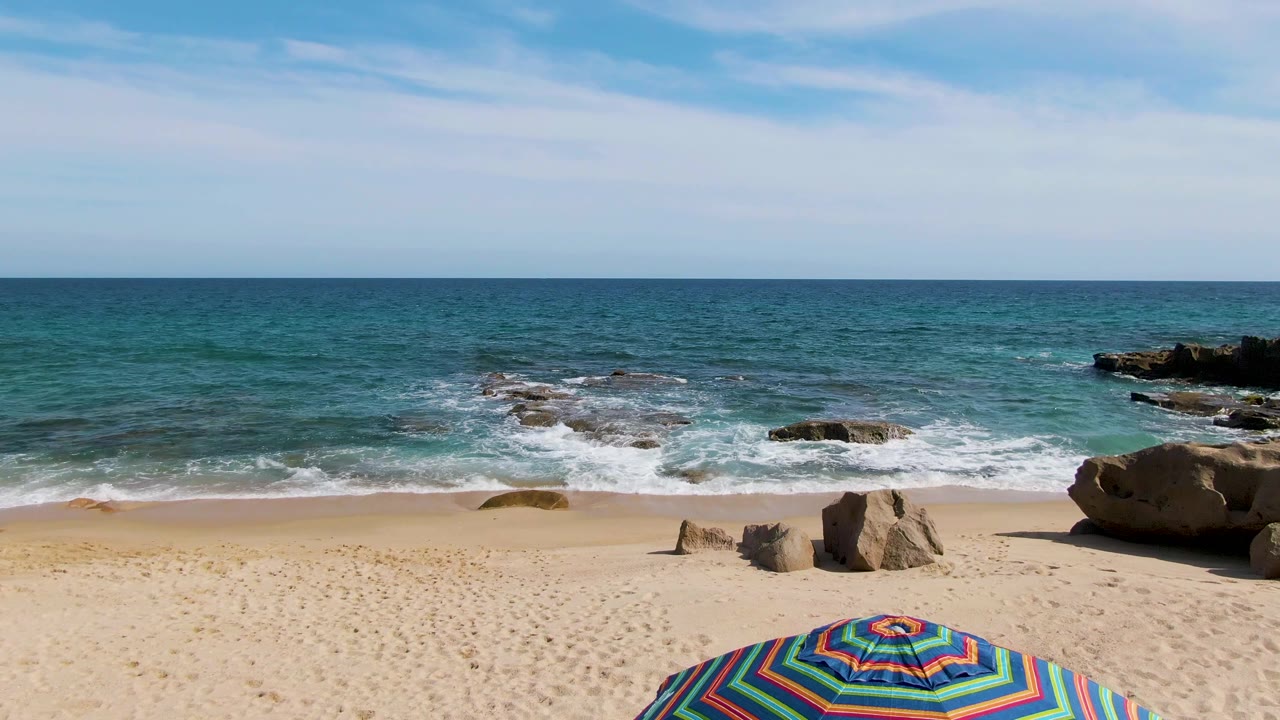 Punta Gorda Beach shoreline with the striped umbrella lower in the frame, scattered rocks on the sand, white surf around offshore rocks, blue-green water, and no visible people.