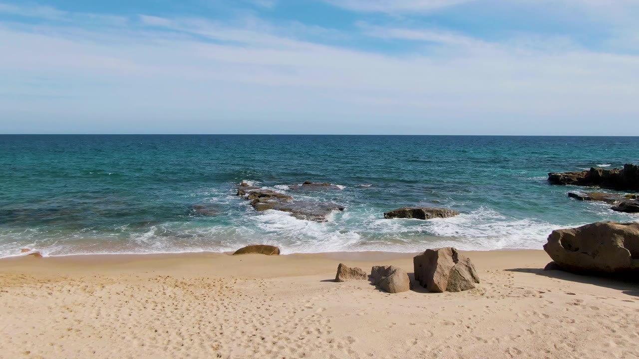Punta Gorda Beach with open sand in the foreground, exposed rocks near the waterline, white foam on the incoming waves, a level horizon, light cloud cover, and no visible people.