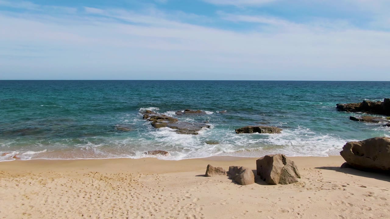 Open sandy beach with blue ocean and rocks under a lightly clouded sky