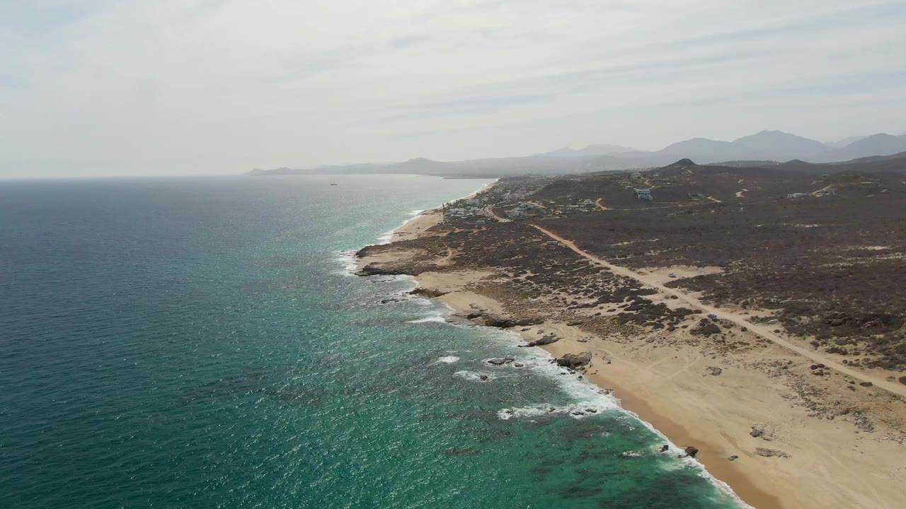 High aerial frame of Punta Gorda Beach, showing white foam advancing along the rocky edge, a broad sand strip, turquoise shallows, dark scrubland, and no visible people.