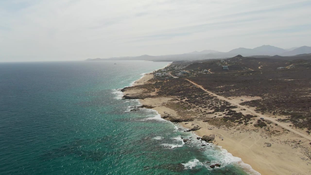 Opening aerial view of Punta Gorda Beach in Baja California Sur, with turquoise water on the left, surf along sand and rocks, a dirt road through scrubland, distant mountains, and no visible people.