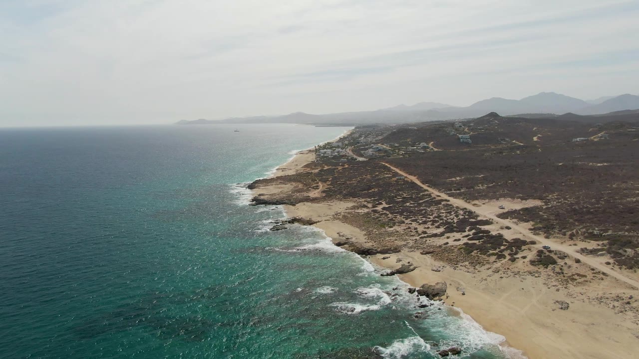 Aerial frame over Punta Gorda Beach showing white waves along the sand, exposed rocks near the waterline, a pale coastal road crossing dark desert vegetation, and no visible people.