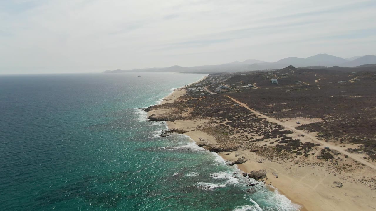 Punta Gorda Beach aerial frame with blue-green water taking more of the left side, foamy surf tracing the beach, desert scrub and road on the right, distant hills, and no visible people.