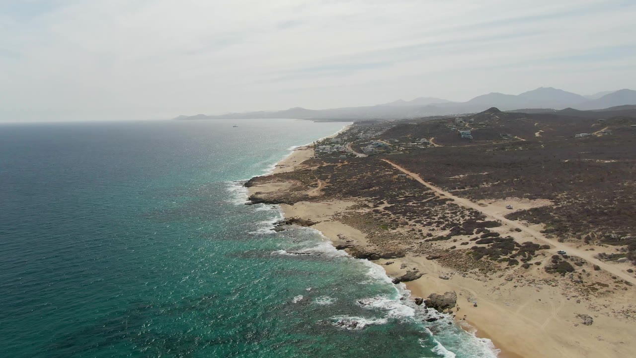 Mid-clip aerial view of Punta Gorda Beach with the ocean edge shifted farther left, surf breaking around nearshore rocks, sandy beach below, scrubland and a road inland, and no visible people.