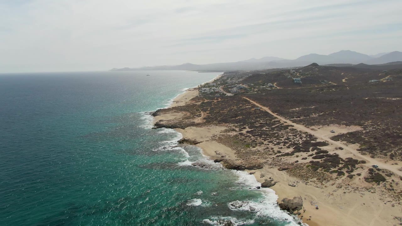 Aerial frame over Punta Gorda Beach, Baja California Sur, with turquoise water on the left, a rocky sandy shoreline on the right, distant mountains, and no visible people.