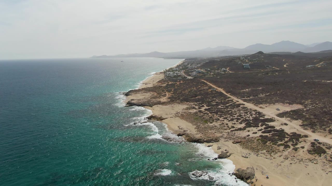 Aerial frame at Punta Gorda Beach showing waves breaking against rocky sand, the dirt road running inland from the shore, hazy mountains beyond, and no visible people.