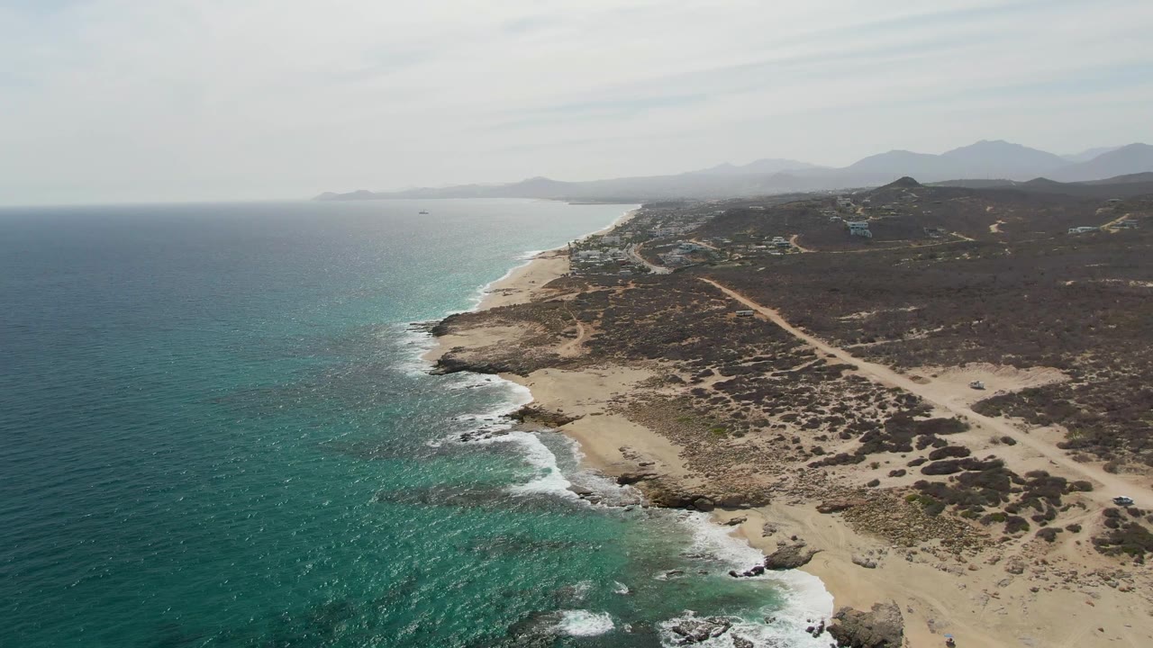 Drone view over Punta Gorda Beach with blue-green ocean water, pale sandy coves, dark scrubland, a road across the headland, distant hills, and no visible people.