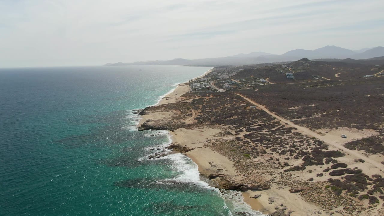 Aerial view of Punta Gorda Beach in Baja California Sur with turquoise water on the left, surf along a rocky sandy shore, dirt roads inland, distant hills, and no visible people.