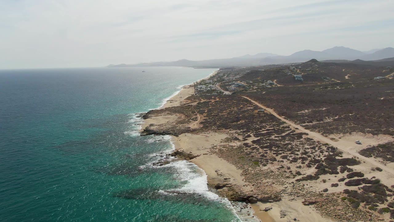 Punta Gorda Beach coastline from above, showing a broader band of turquoise water, white foam against the rocks, sandy shore, dirt tracks, distant hills, and no visible people.