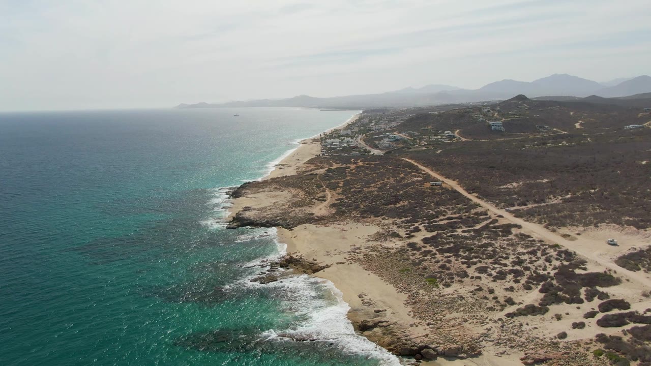 Aerial beach view with ocean on the left, rocky surf line, inland scrub, and dirt road.
