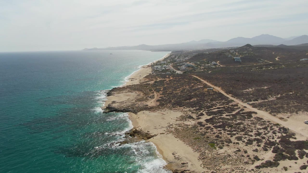 Aerial view over Punta Gorda Beach in Baja California Sur with turquoise ocean at left, rocky shoreline below, sandy beach, inland dirt road, distant mountains, and no visible people.