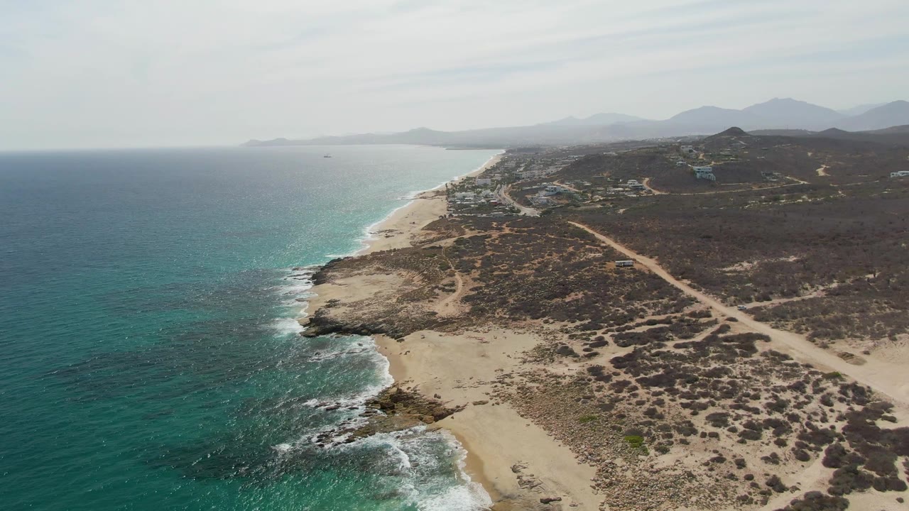 Aerial frame of Punta Gorda Beach showing a slightly closer rocky point with white surf, teal water, pale sand, scrub-covered terrain, a dirt road, distant mountains, and no visible people.