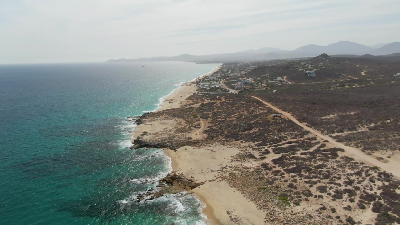 Aerial view at Punta Gorda Beach with the shoreline running diagonally through frame, turquoise ocean to the left, sand and rocks below, inland dirt road, low hills, distant mountains, and no visible people.