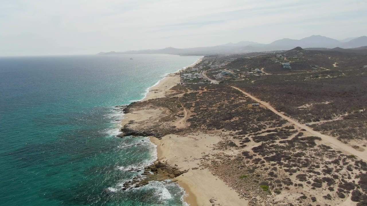 Coastal aerial with open ocean, sandy beach, rocky foreground, scrubland, and mountain horizon.