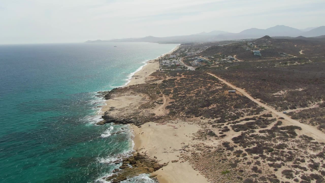 Aerial coastline with turquoise ocean, sandy beach, dry scrubland, dirt roads, and distant mountains.