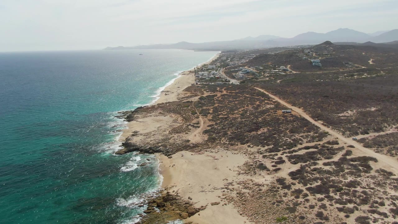 Aerial frame from Punta Gorda Beach showing small white waves along the rocky sand edge, clear turquoise water, dry vegetation, dirt tracks, hazy mountains, and no visible people.