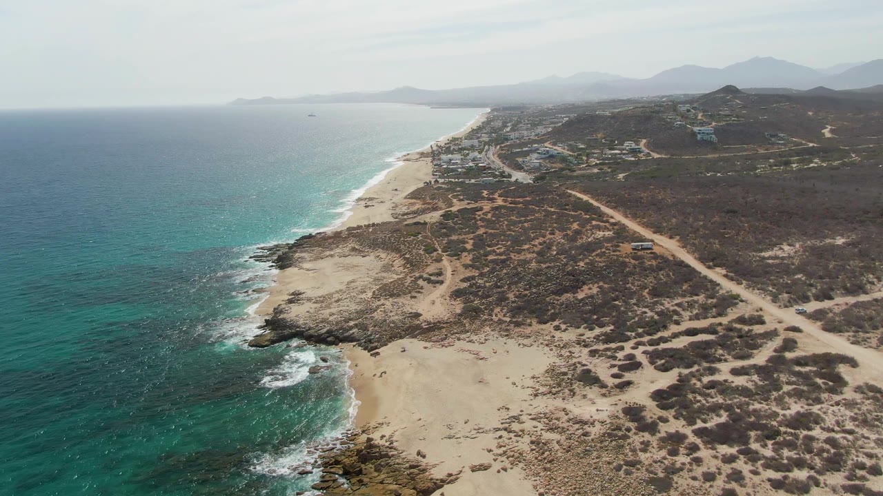 Punta Gorda Beach aerial frame with blue-green ocean filling the left side, a curved sandy and rocky shoreline below, sparse dry vegetation inland, distant hills and mountains, and no visible people.