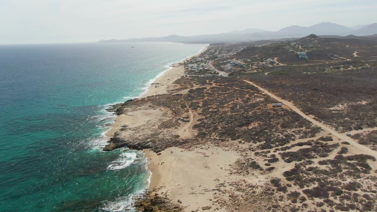 Mid-clip aerial view at Punta Gorda Beach with turquoise water and white wave patches on the left, broad sandy beach at the bottom, dry brush and a dirt road inland, distant mountains, and no visible people.