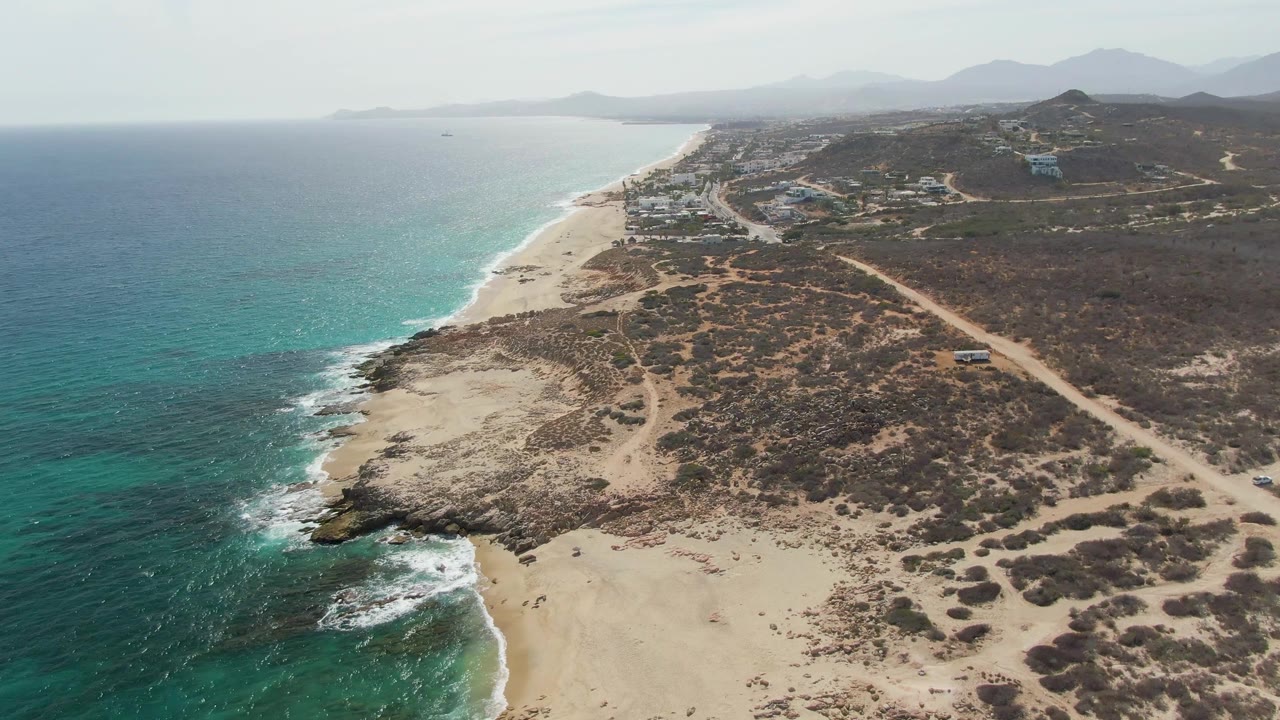 Opening aerial frame over Punta Gorda Beach, Baja California Sur, with turquoise water at left, a sandy shoreline below, dry scrub hills inland, distant mountains, and no visible people.