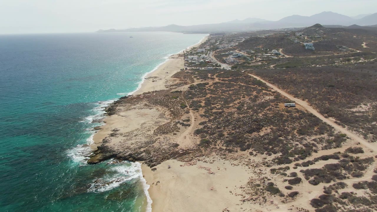 Drone view along a sandy coast with waves, scrub-covered hills, a dirt road, and buildings in the distance.