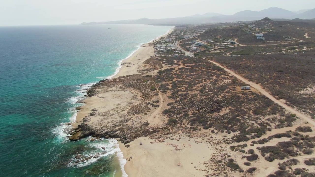 Drone frame over Punta Gorda Beach showing foamy water around shoreline rocks, a broad sandy beach, a dirt road through scrubland, distant development, and no visible people.