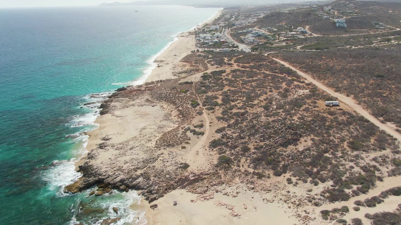 Aerial view at Punta Gorda Beach, Baja California Sur, with turquoise ocean on the left, pale sand and scrub-covered dunes below, a dirt road to the right, and no visible people.