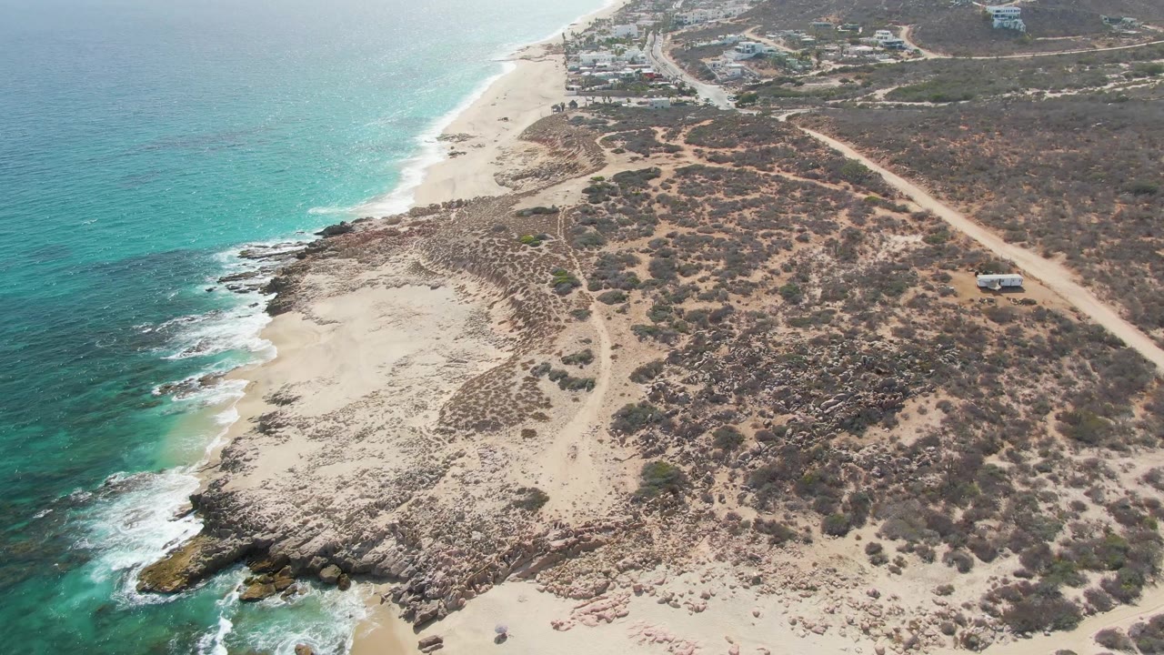 At 0:02, the Punta Gorda Beach coastline fills the frame with clear blue-green water, surf along the rocks, sand and scrub inland, a dirt road at right, a small vehicle, and no visible people.