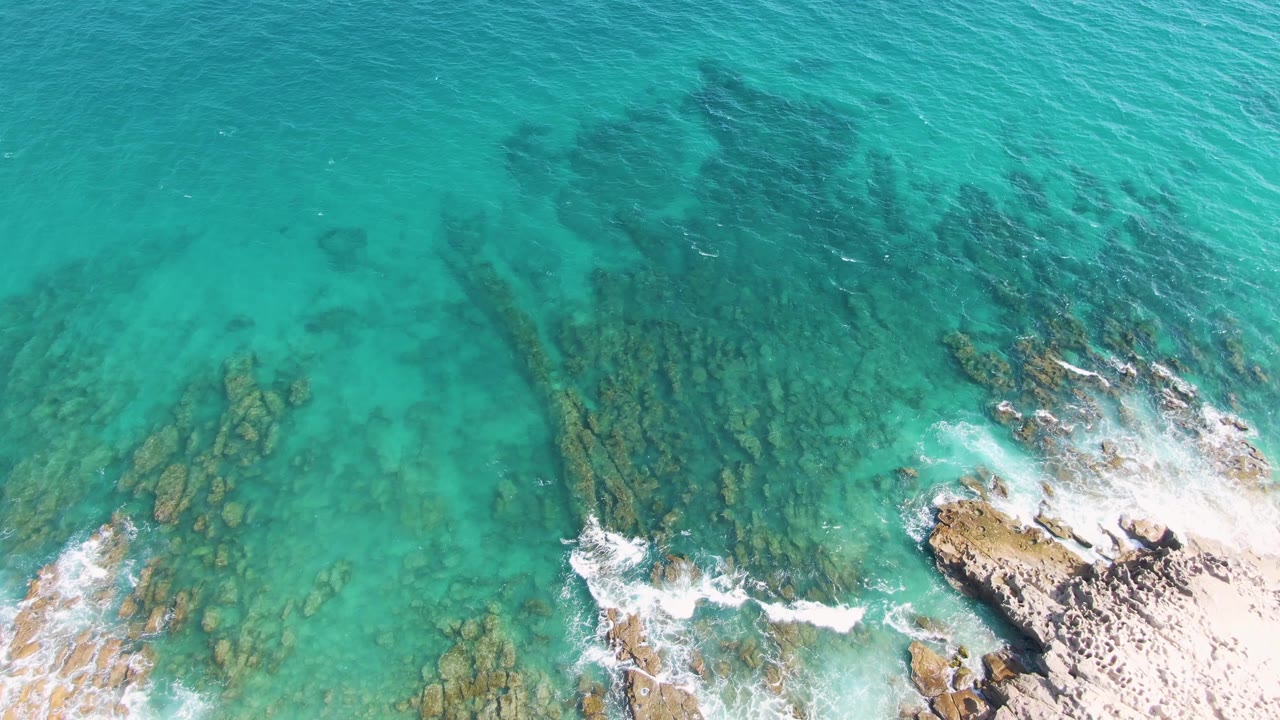 Aerial frame from Punta Gorda Beach with white surf spreading over submerged rock shelves in clear turquoise water and no visible people.